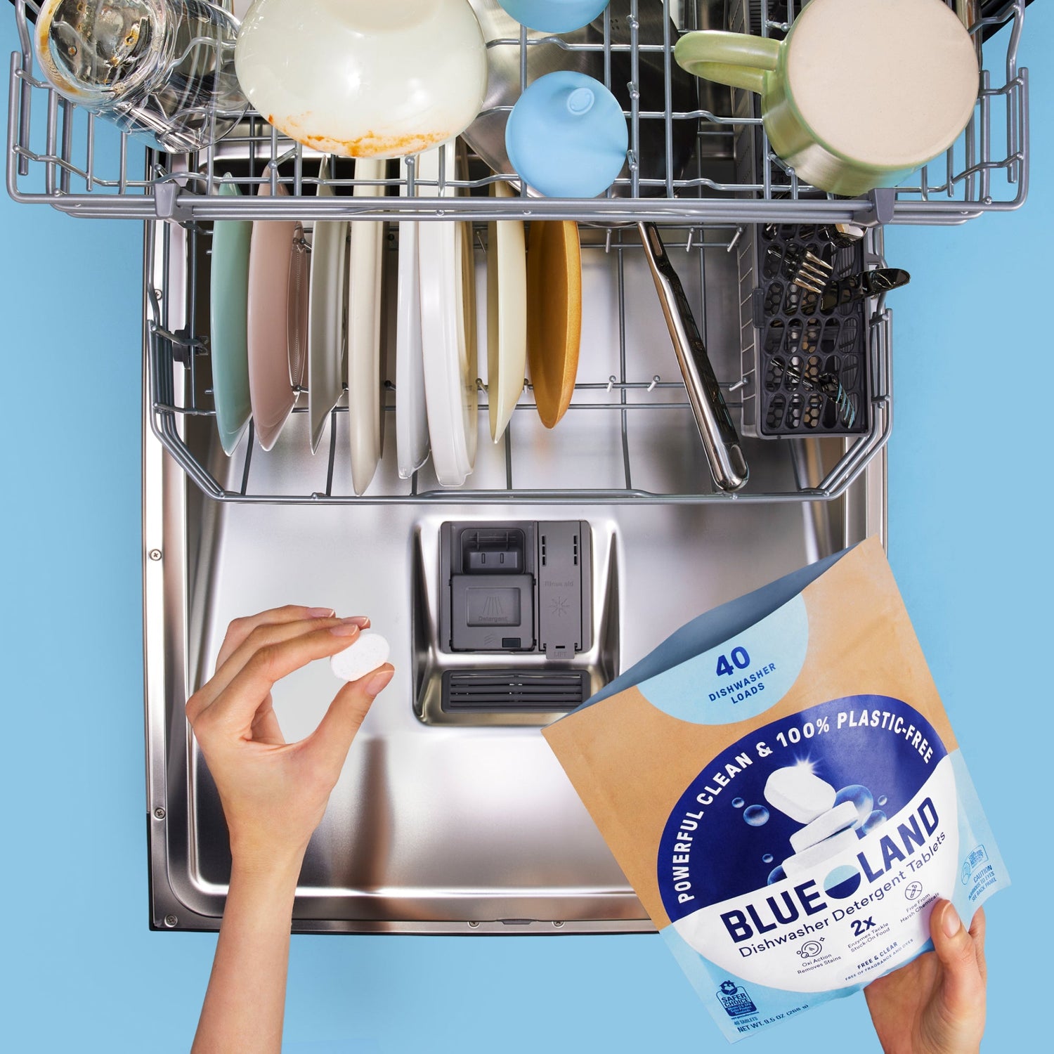 A hand holds a dishwasher tablet above an open dishwasher filled with clean dishes; a Blueland dishwashing detergent tablet package is visible in the foreground.